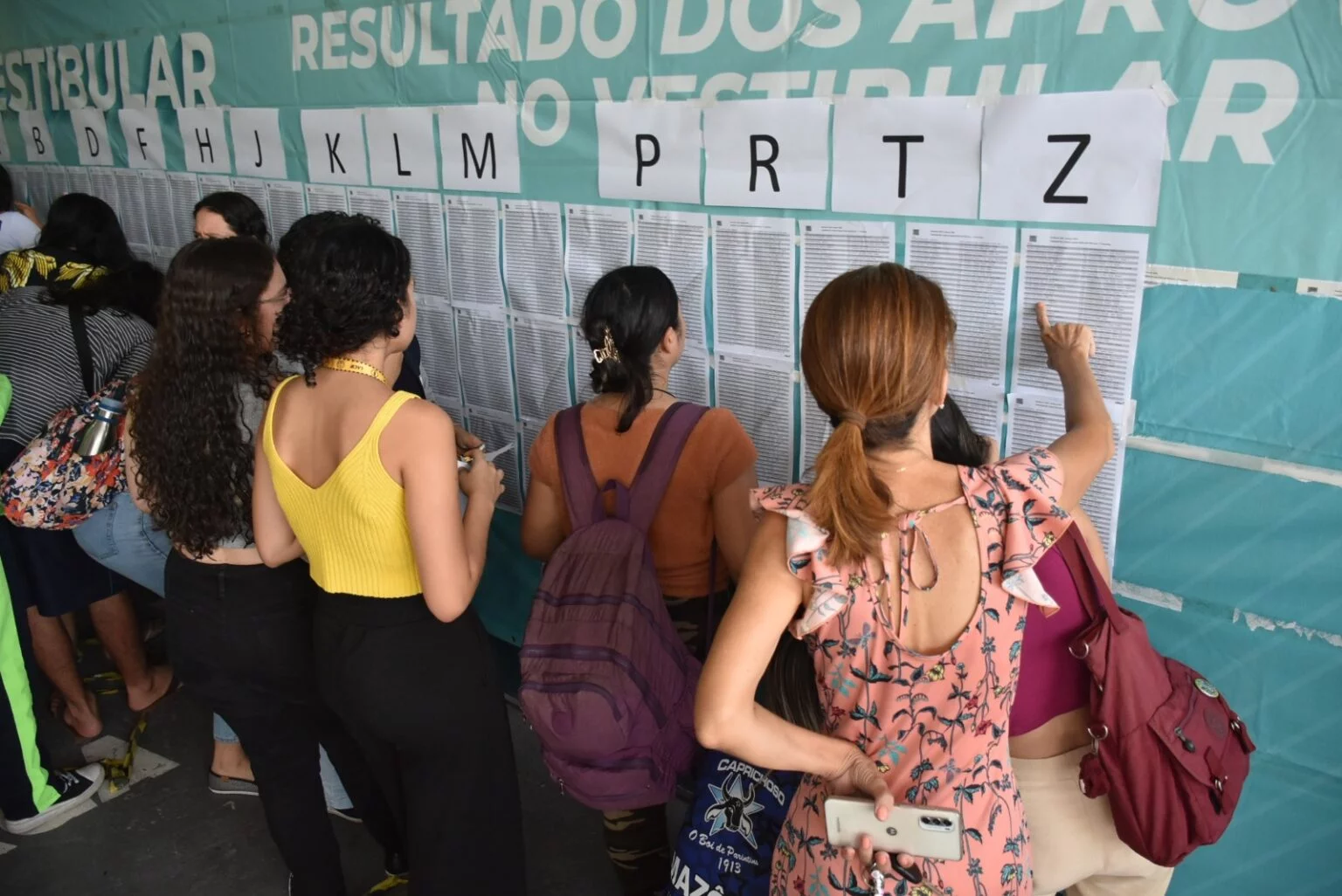 Imagem colorida mostra concorrentes procurando nomes em lista fixadas na parede