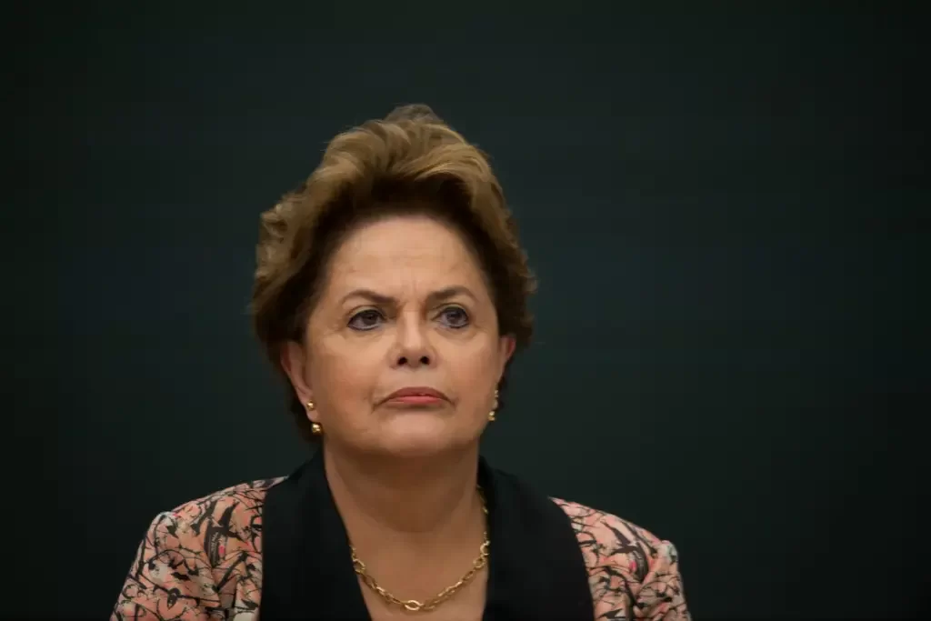 Imagem colorida mostra Dilma Rousseff séria com um fundo escuro